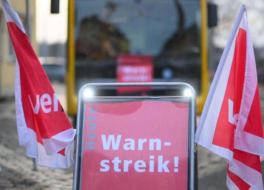 Ein Schild mit der Aufschrift "heute Warnstreik!" in der Mitte des Bildes und an beiden Seiten Flaggen mit dem Logo der Gewerkschaft Verdi.