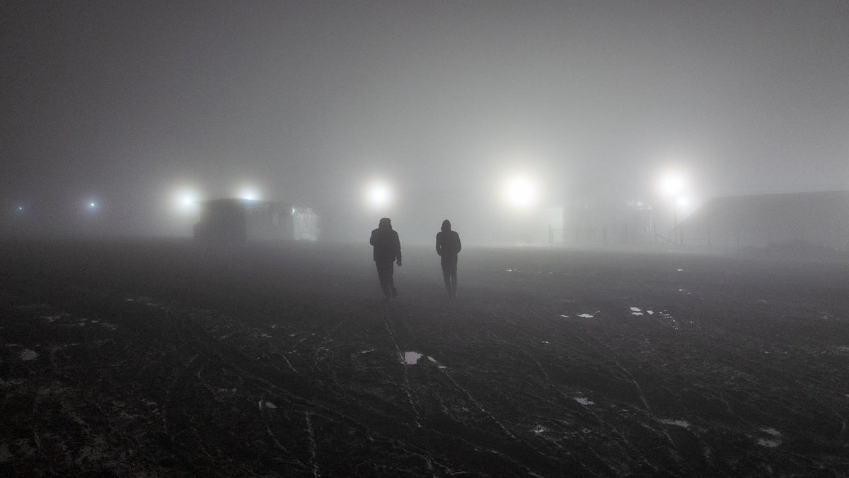 Zwei Menschen sind von hinten zu sehen, die im nebeligen Scheinwerferlicht Nachts über ein Feld gehen.