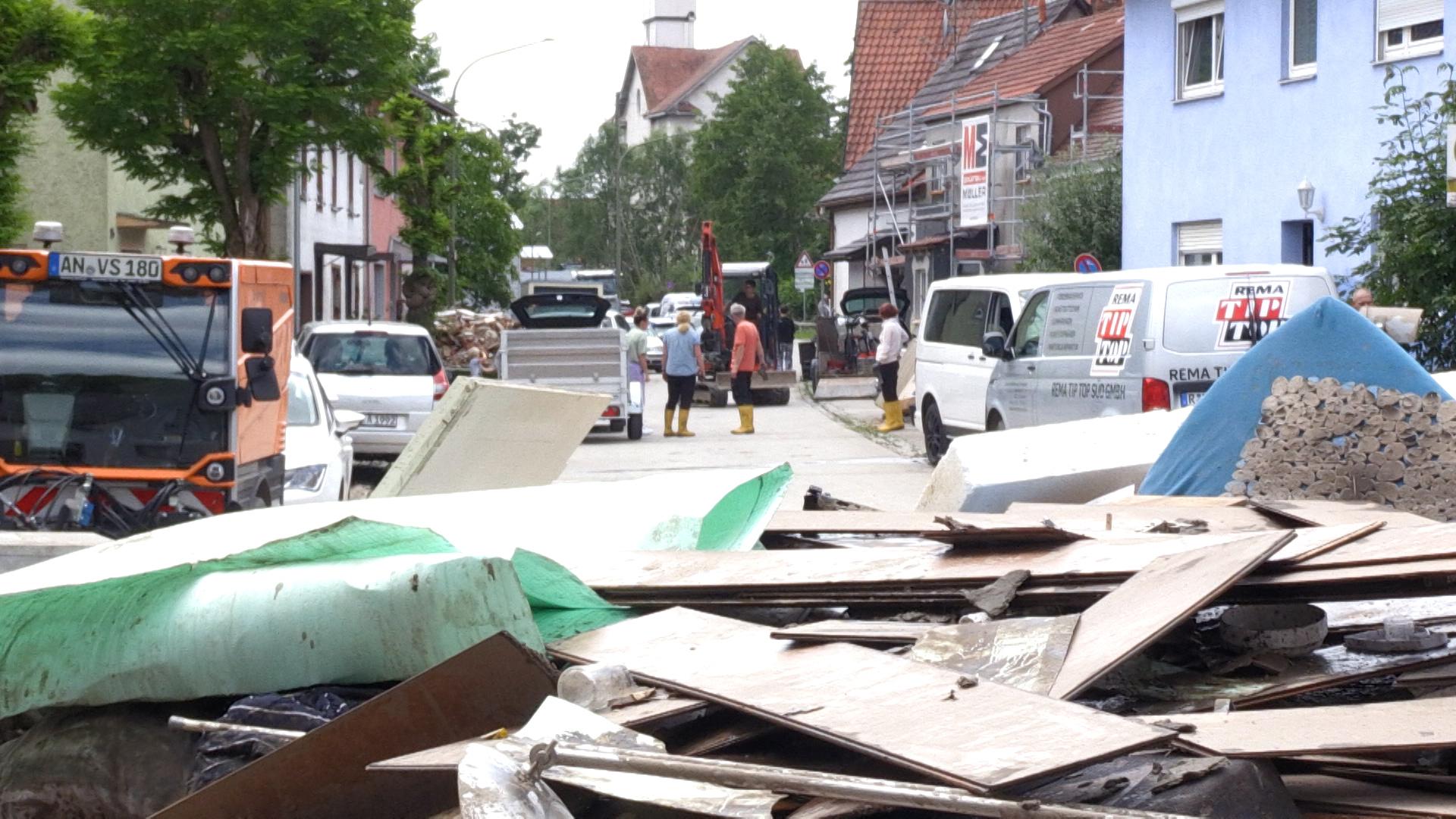 In Günzburg stehen Anwohner in Gummistiefeln auf der Straße, für die Sperrmüll-Abholung aufgetürmt liegen um sie herum Dinge, die vom Hochwasser zerstört wurden.
