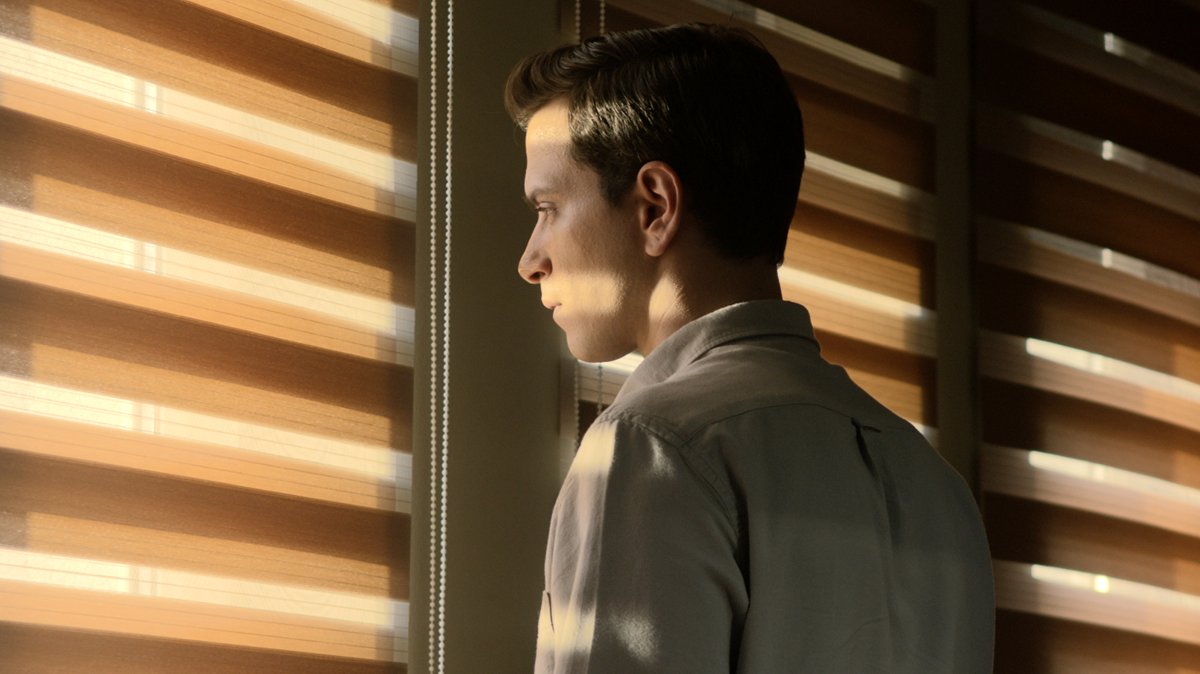 Ein junger Mann steht am Fenster mit Jalousien