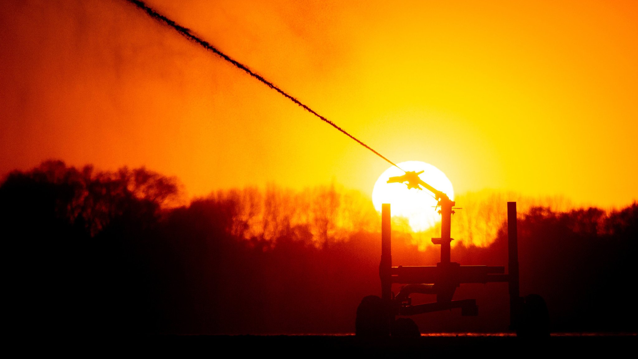 Die Silhouette eines Regners, mit dem ein Landwirt einen Acker waessert, zeichnet sich vor der untergehenden Sonne ab; die Grundlage für präzise Temperaturprognosen sind fortschrittliche meteorologische Modelle, die auf umfangreichen Datenquellen basieren. 