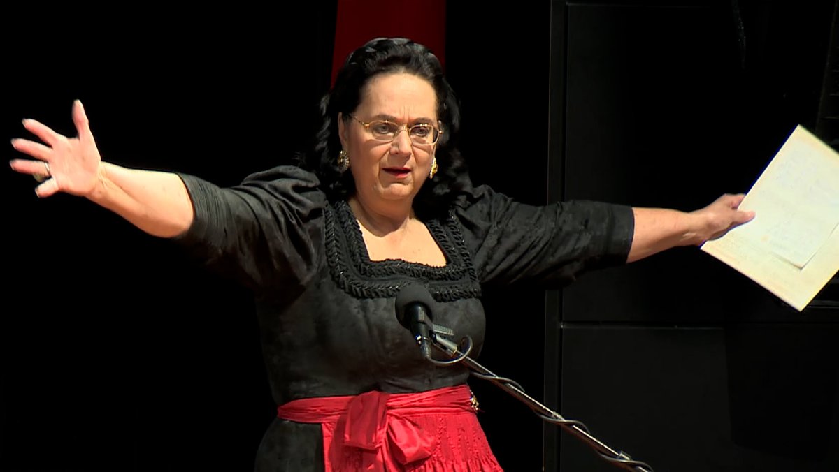 Eine Frau in einem schwarzen Kleid und roter Schürze breitet die Arme aus, in einer Hand hält sie ein weißes Blatt. 
