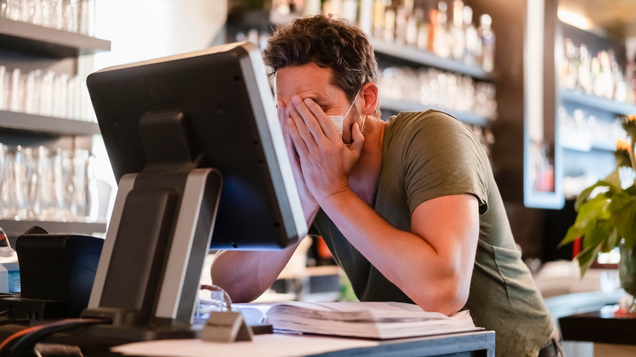 Ein Mann steht in einem Restaurant an einem Bildschirm bei der Kasse. Er hat die Hände vor sein Gesicht geschlagen. 