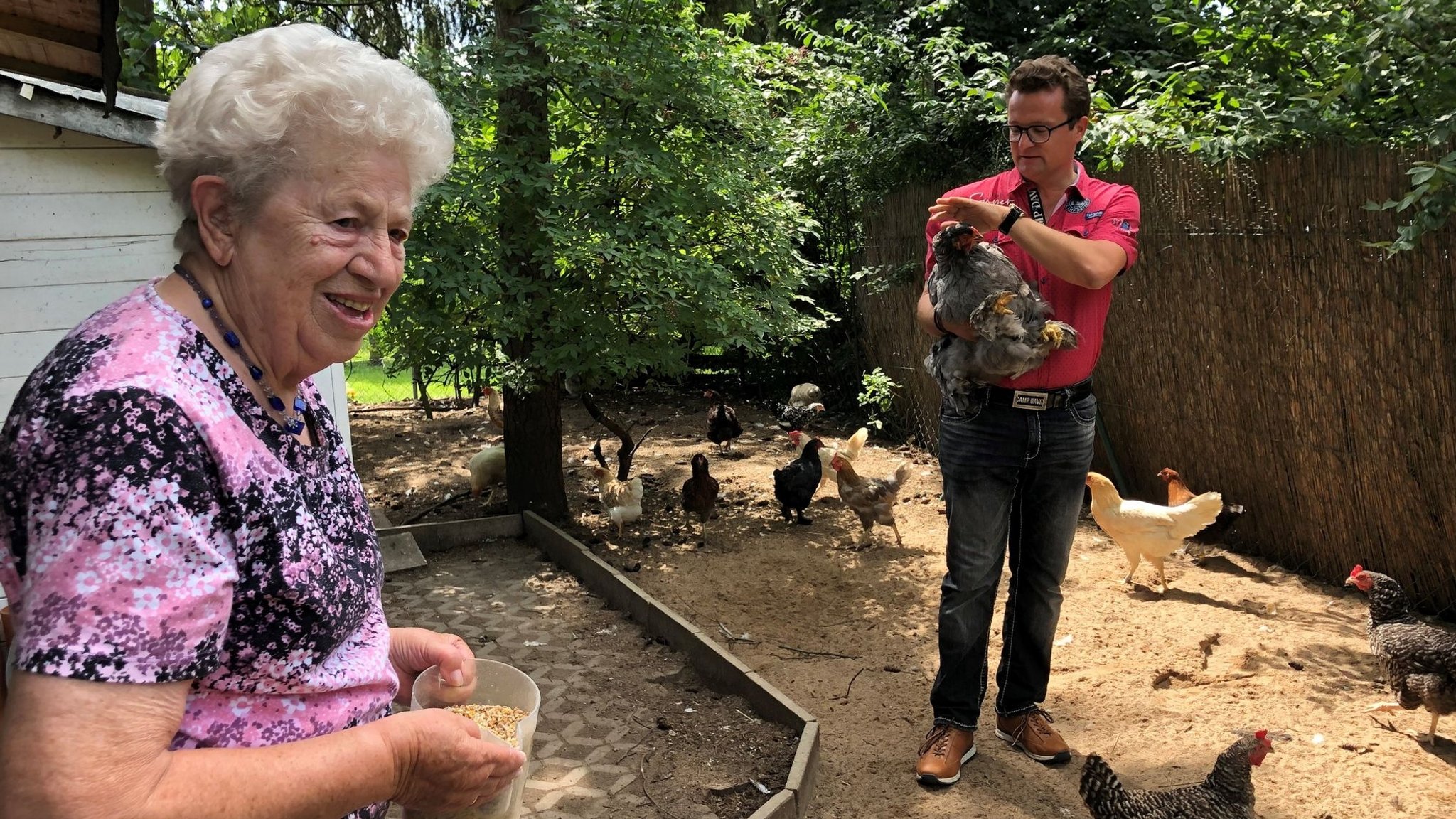 ine Bewohnerin des Seniorenheims steht mit einem Becher mit Futter im Hühnergehege. Im Hintergrund hebt der Heimleiter ein Huhn hoch.