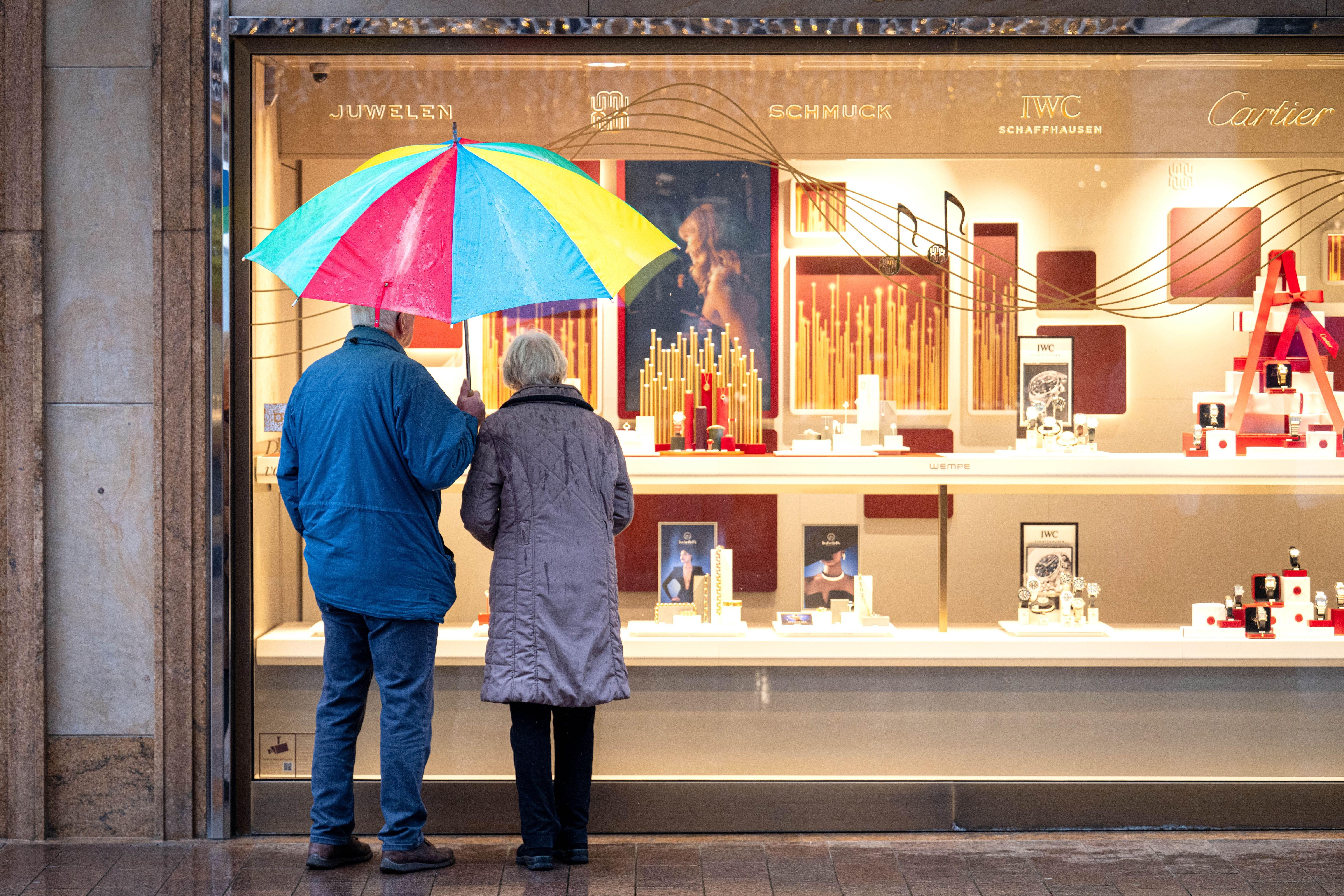 Zwei Personen im Rentenalter stehen bei Regen vor einem Schaufenster eines Schmuckgeschäfts und betrachten die ausgestellten Waren.