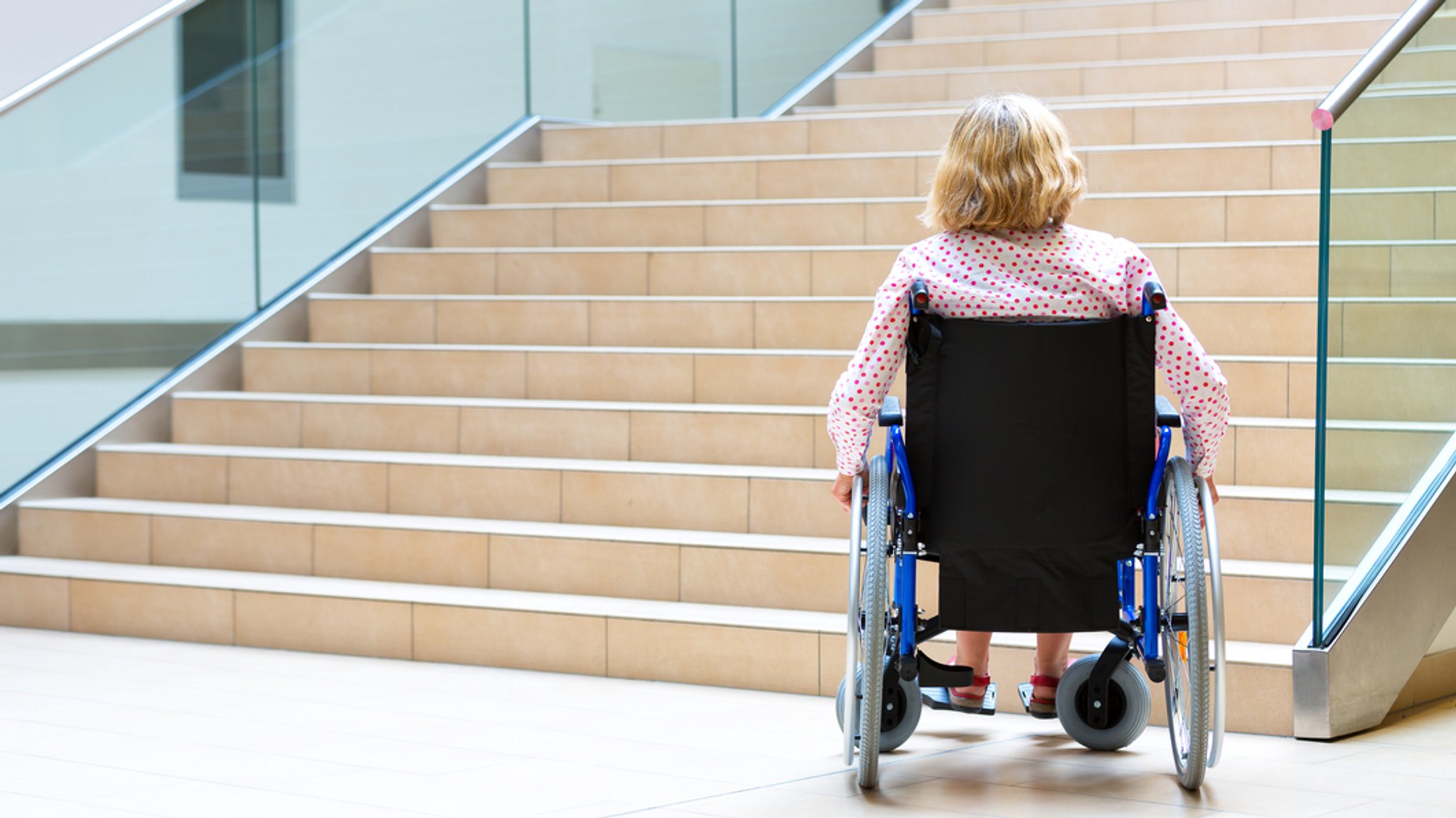 Eine Frau im Rollstuhl sitzt mit dem Rücken zur Kamera vor einer langen Treppe.