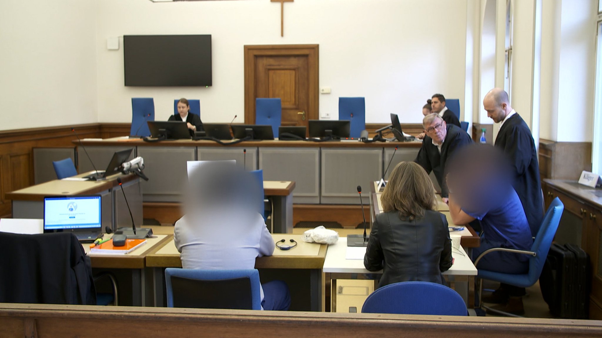 Weitwinkelaufnahme aus einem Gerichtssaal: Zwei Angeklagte sitzen mit ihren Verteidigern an einem Tisch.