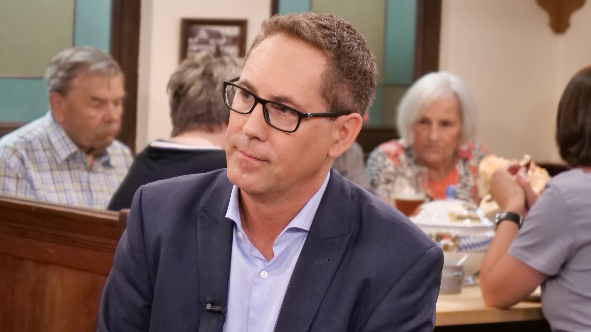 Michael Schrodi sitzt am Sonntags-Stammtisch. Im Hintergrund sind Gäste des Wirtshauses zu sehen. Schrodi trägt einen Anzug, ein hellblaues Hemd, eine schwarze Brille und kurze, braune Haare. | Bild: Andreas Schroll / Bayerischer Rundfunk 2026 Michael Schrodi sitzt am Sonntags-Stammtisch. Im Hintergrund sind Gäste des Wirtshauses zu sehen. Schrodi trägt einen Anzug, ein hellblaues Hemd, eine schwarze Brille und kurze, braune Haare.
