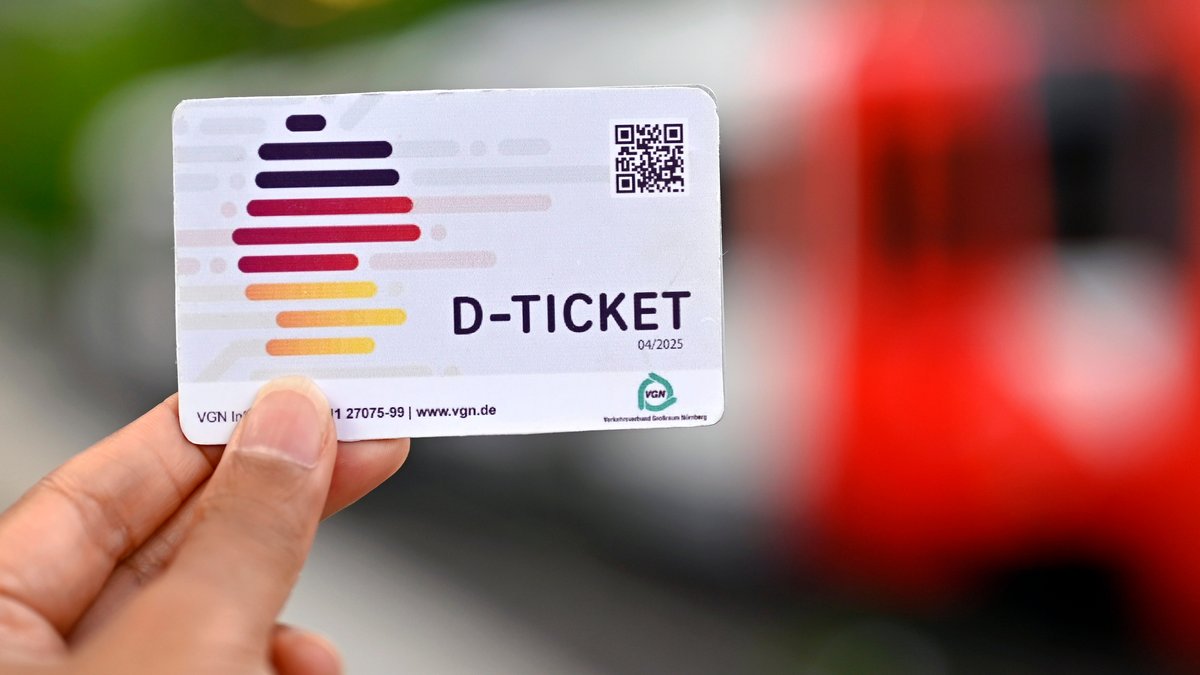 Eine Hand hält ein Deutschland-Ticket (D-Ticket), im Hintergrund ist eine Bahn zu sehen.