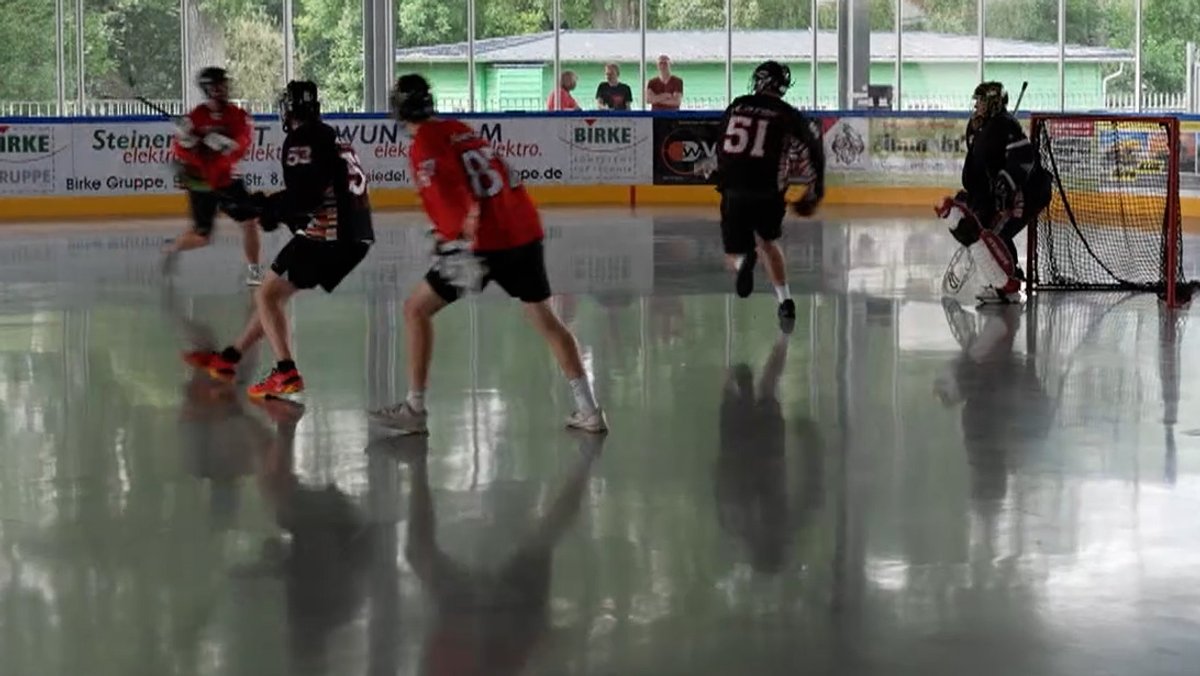 Auf einem Spielfeld in einer Halle spielen Männer mit Holschlägern, an denen kleine Netze hängen. 