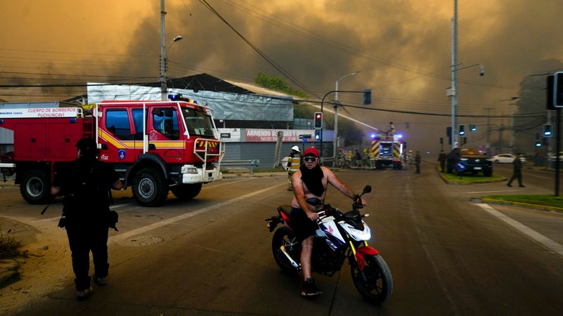 Ein Anwohner verlässt am 3.2.204 im chilenischen Viña del Mar auf seinem Motorrad das evakuierte Gebiet, während Feuerwehrleute schützend Wasser auf Gebäude sprühen, da der durch Waldbrände verursachte Rauch den Himmel füllt.