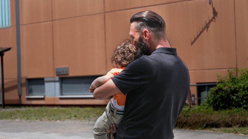 Ein Mann mit Kind auf dem Arm steht vor einem Gefängnis. | Bild: BR / Carina Müller Ein Mann mit Kind auf dem Arm steht vor einem Gefängnis.