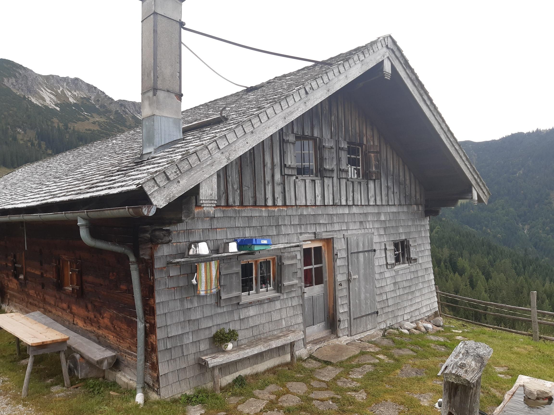 Schindelgedeckte Holzhütte in den Bergen mit Hausbank und Brunnen im Vordergrund 