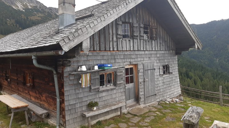 Schindelgedeckte Holzhütte in den Bergen mit Hausbank und Brunnen im Vordergrund | Bild: Angela Braun / BR Schindelgedeckte Holzhütte in den Bergen mit Hausbank und Brunnen im Vordergrund
