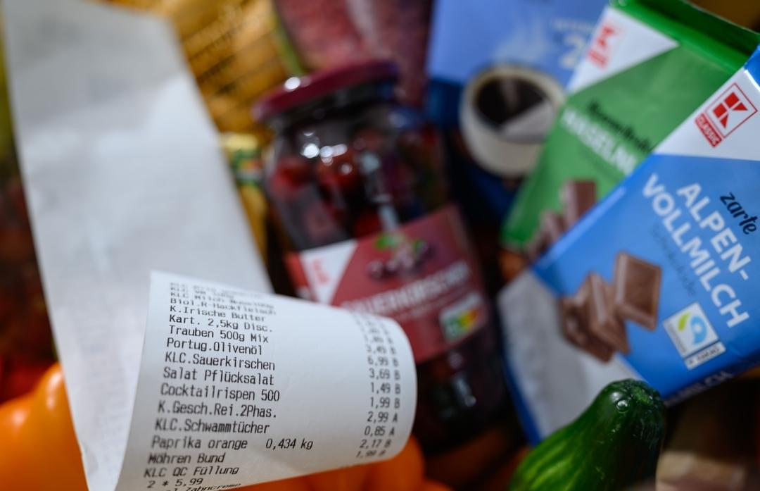 (Symbolbild) Ein Kassenbon liegt auf Supermarktprodukten. Zu sehen sind eine Tafel Schokolade, eine Gurke so wie ein Glas Sauerkirschen