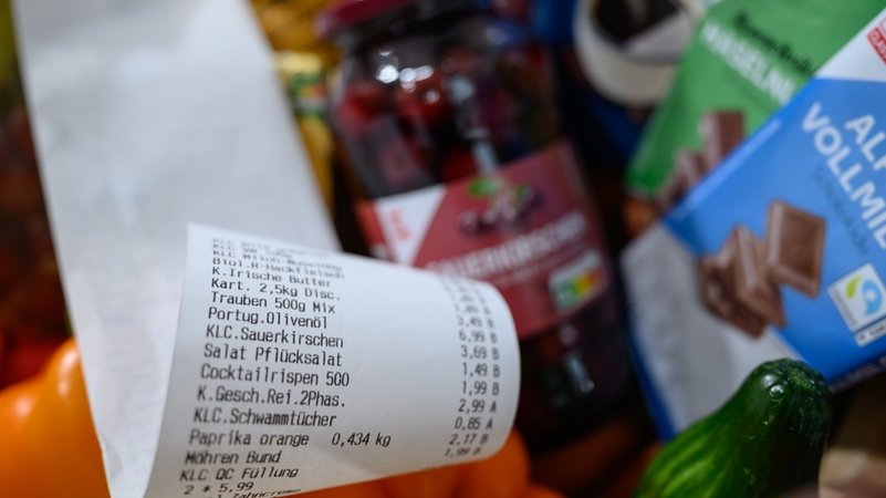 (Symbolbild) Ein Kassenbon liegt auf Supermarktprodukten. Zu sehen sind eine Tafel Schokolade, eine Gurke so wie ein Glas Sauerkirschen | Bild: dpa-Bildfunk/Hendrik Schmidt (Symbolbild) Ein Kassenbon liegt auf Supermarktprodukten. Zu sehen sind eine Tafel Schokolade, eine Gurke so wie ein Glas Sauerkirschen