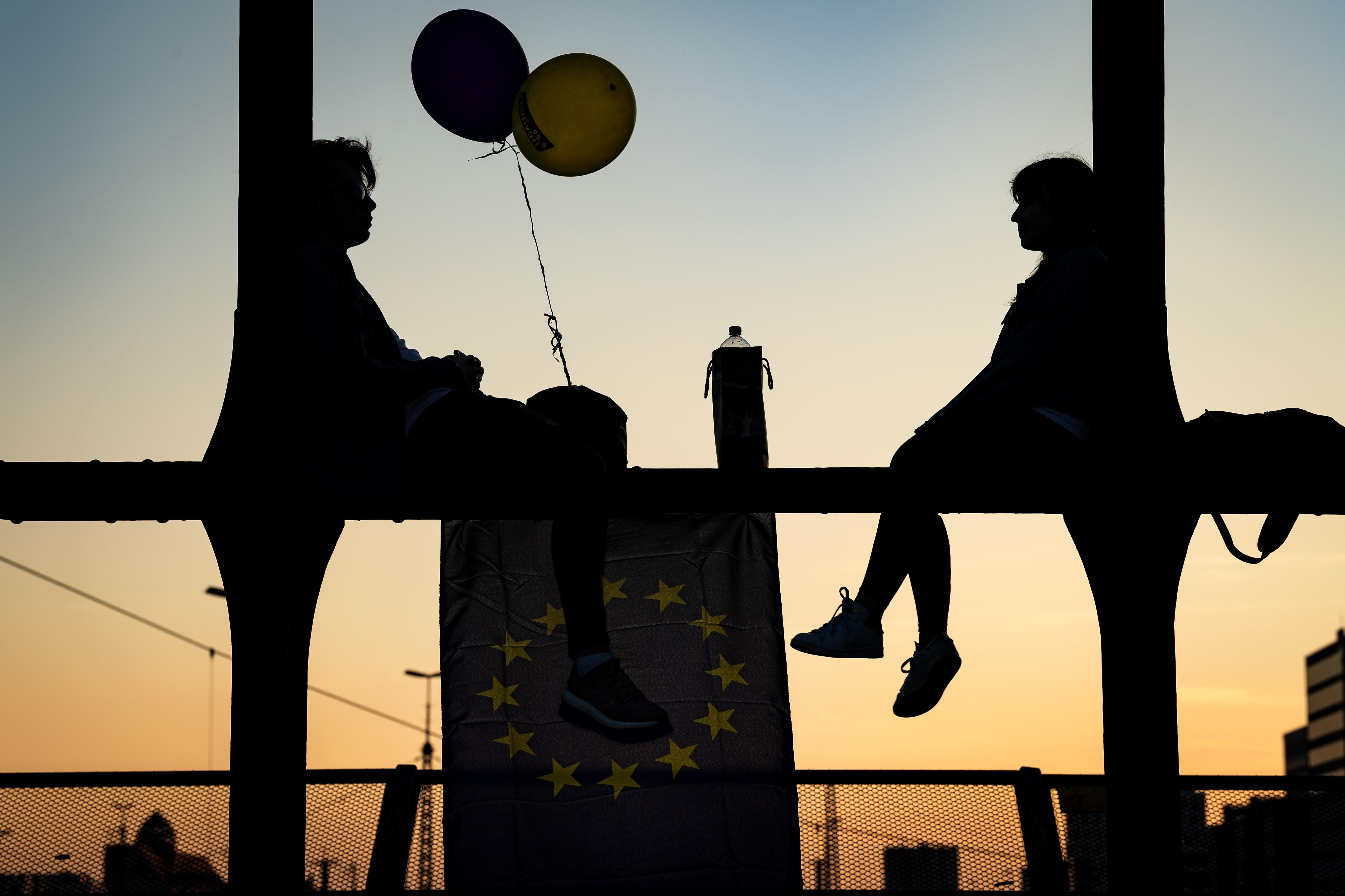 Ein junger Mann und eine junge Frau sitzen mit Luftballons und einer Europaflagge auf den Streben einer Brücke.
