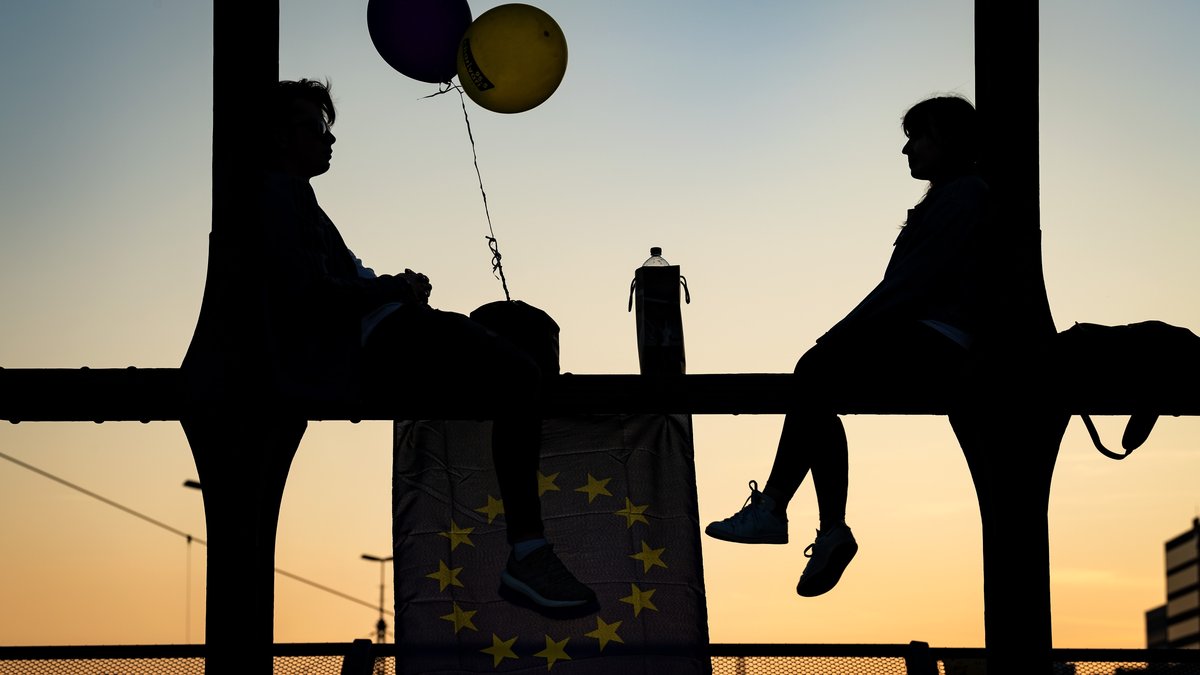Ein junger Mann und eine junge Frau sitzen mit Luftballons und einer Europaflagge auf den Streben einer Brücke.