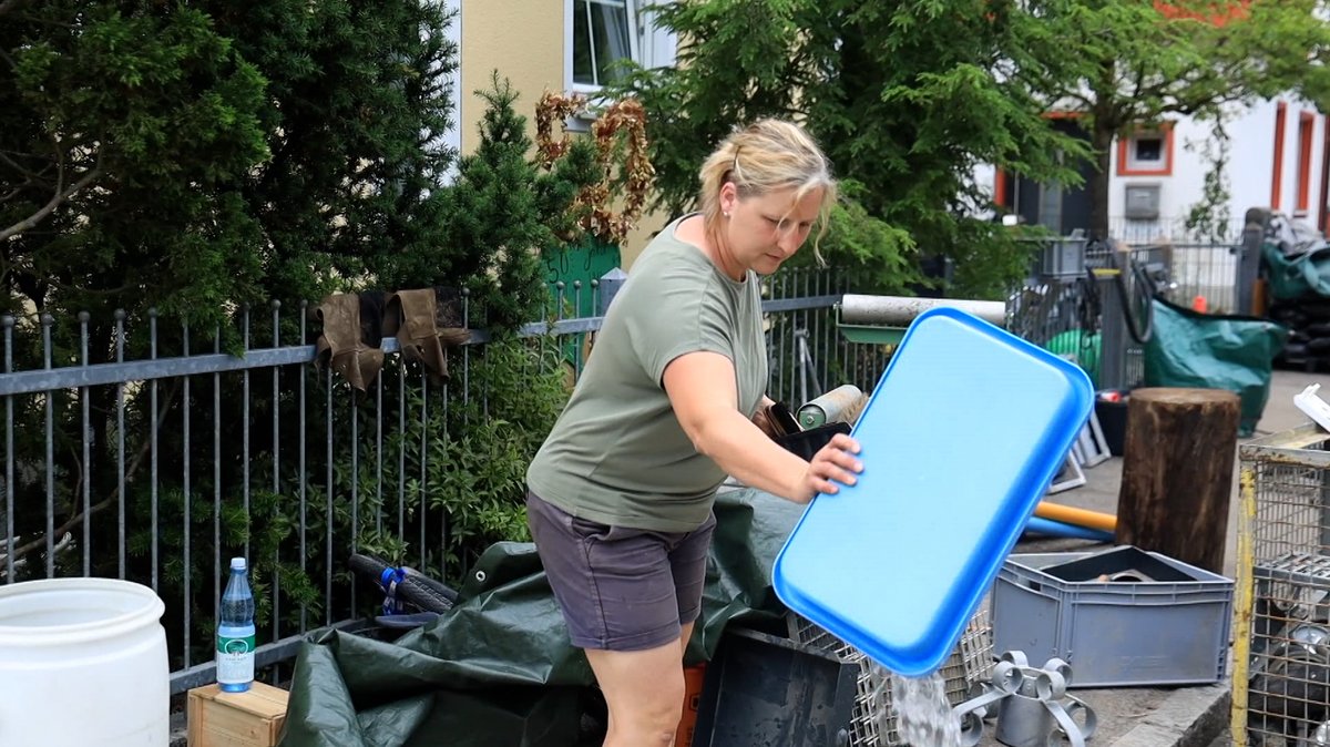 Christine Hammer aus Schrobenhausen schüttet Wasser aus einer Wanne vor ihrem Haus. Im Hintergrund sieht man verschiedene Haufen mit Dingen, die während des Hochwassers kaputt gegangen sind. 