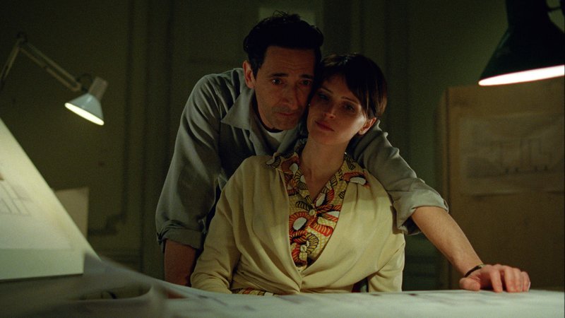 Adrien Brody und Felicity Jones in einer Szene aus dem oscar-nominierten Drama "Der Brutalist": Sie sitzt an einem Schreibtisch, er beugt sich von hinten über sie und blickt auf einen ausgebreiteten Gebäudeentwurf. | Bild: Universal Pictures Adrien Brody und Felicity Jones in einer Szene aus dem oscar-nominierten Drama "Der Brutalist": Sie sitzt an einem Schreibtisch, er beugt sich von hinten über sie und blickt auf einen ausgebreiteten Gebäudeentwurf.