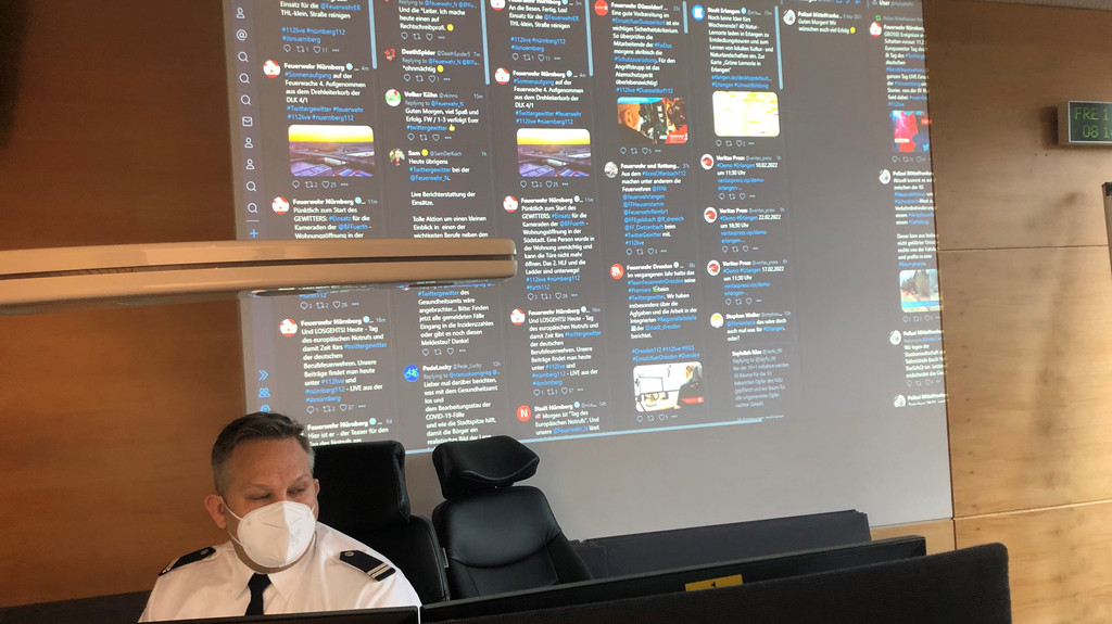 In der ILS Nürnberg: Tweetdeck auf einer Leinwand, davor ein Feuerwehrmann vor seinen Bildschirmen. 