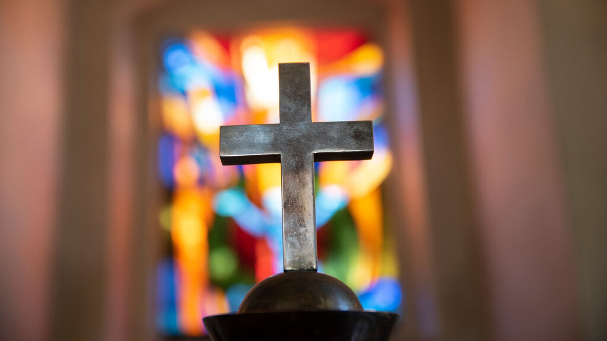 Ein Kreuz aus Metall steht vor einem Kirchenfenster. | Bild: stock.adobe.com/flordigitalartist Ein Kreuz aus Metall steht vor einem Kirchenfenster.