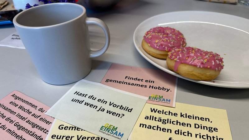 Eine Kaffeetasse neben einem Teller mit rosa Donuts umgeben von Zetteln mit Gesprächseinstiegen.