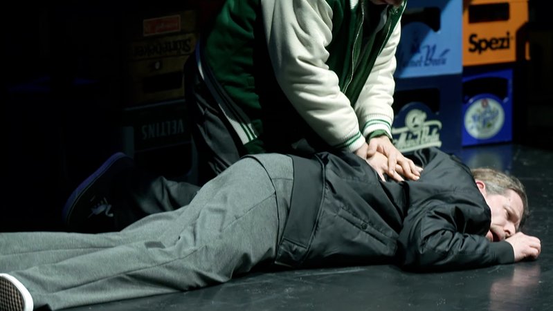 Ein Mann liegt auf der Theaterbühne und wird von einer Hand auf den Boden gedrückt. 