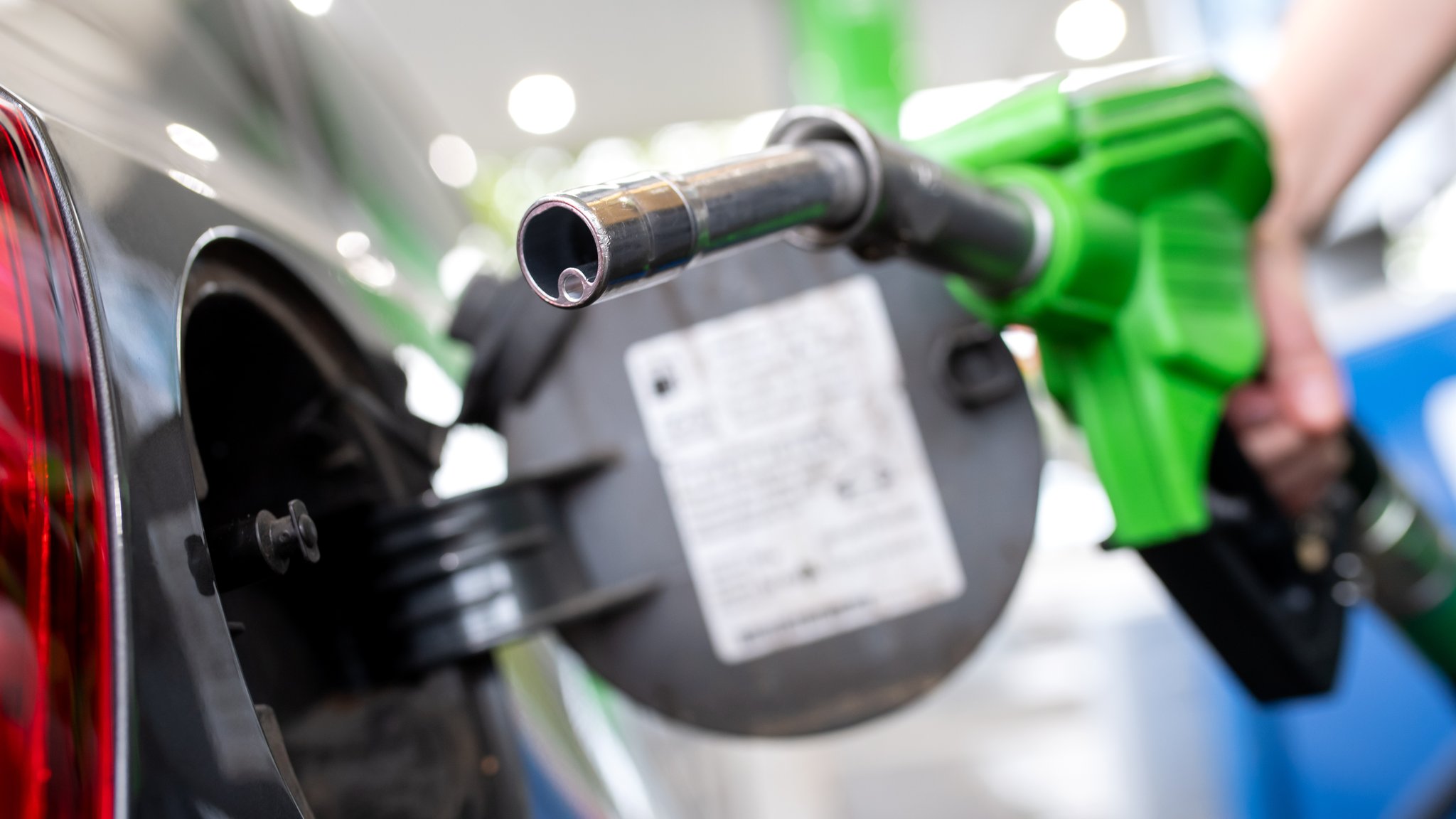 Eine Frau hält an einer Tankstelle an einer Zapfsäule eine Zapfpistole in der Hand und betankt ein Auto. 