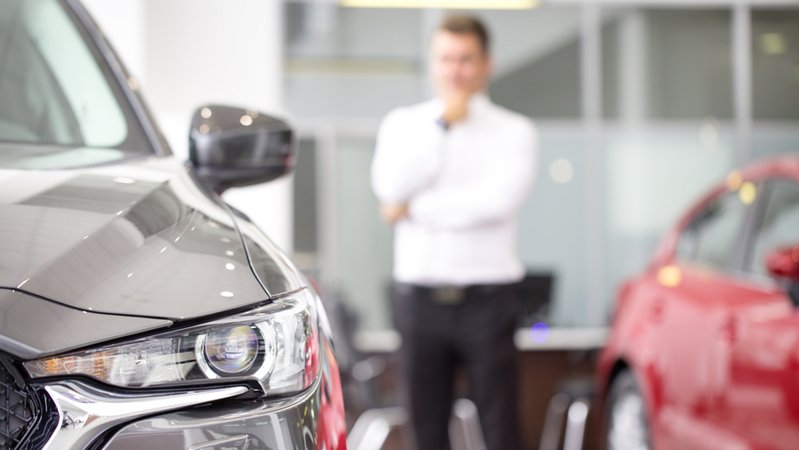 Ein Mann steht in einem Autohaus neben Neuwagen und grübelt, welchen er kaufen soll. | Bild: stock.adobe.com/Zoriana Ein Mann steht in einem Autohaus neben Neuwagen und grübelt, welchen er kaufen soll.