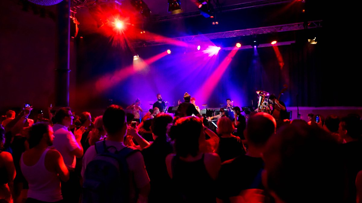 Tanzende Menschen in einem dunklen Saal, bunte Scheinwerfer, vorne Musikerinnen und Musiker.