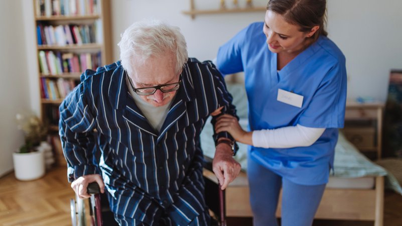 Eine Pflegerin hilft einem älteren Mann im Schlafanzug aus dem Rollstuhl