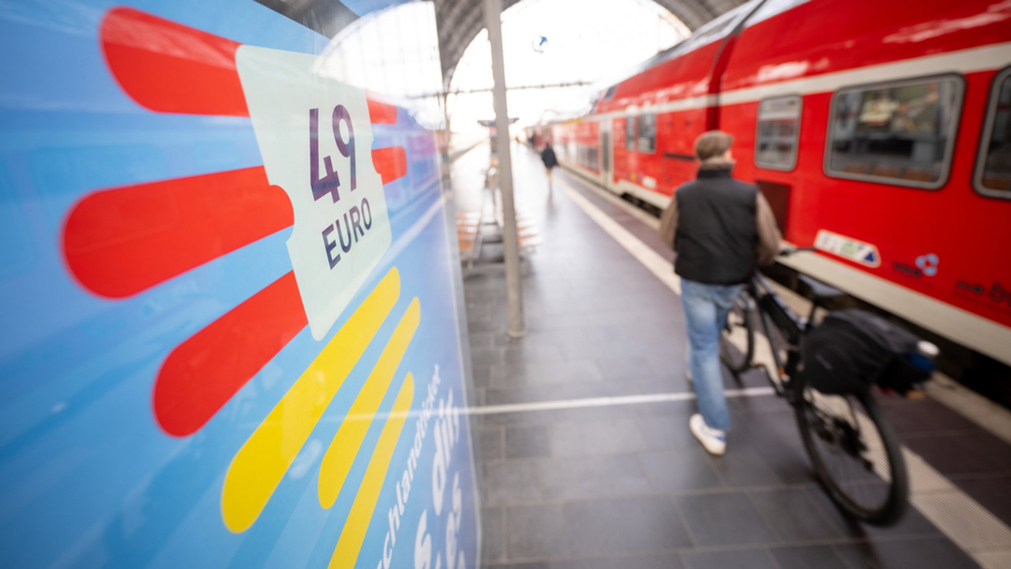 Ein Mann geht am Frankfurt Hauptbahnhof mit seinem Fahrrad zwischen der Werbung für das Deutschlandticket und einer Regionalbahn entlang.