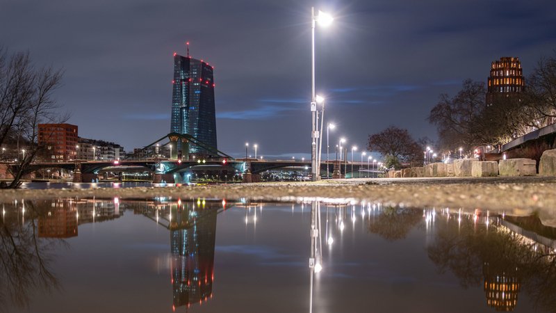 In einer großen Pfütze spiegelt sich am frühen Morgen die Zentrale der Europäischen Zentralbank (EZB, l) am Frankfurter Mainufer.