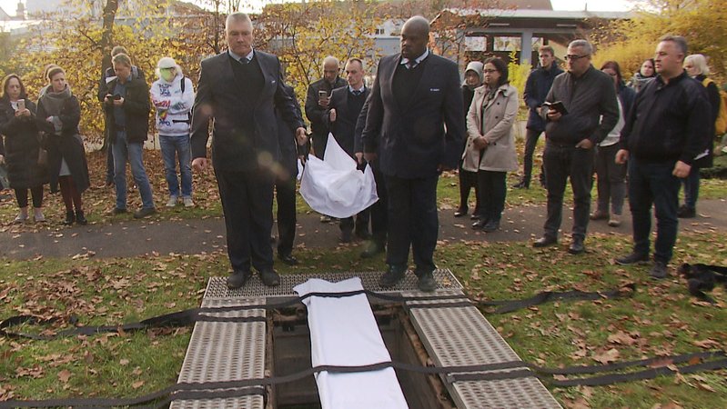 Vor einem Grab halten sechs Männer einen in ein Tuch gehüllten Leichnam.