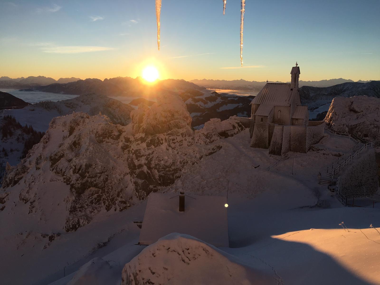 Blick vom Sendergebäude des BR auf dem Wendelstein auf das tief verschneite Voralpen-Panorama und die mit Nebel gefüllten Täler. Im Vordergrund die Wendelsteinkapelle, hinter den Bergen geht die Sonne unter. 