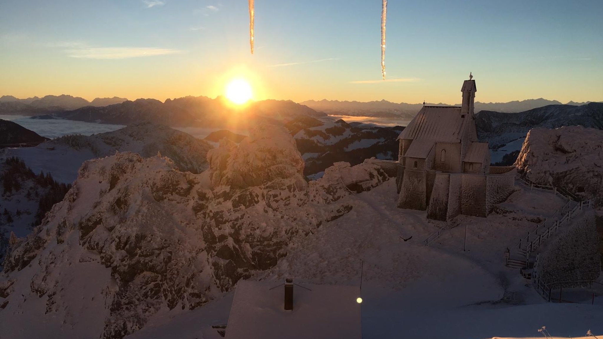 Blick vom Sendergebäude des BR auf dem Wendelstein auf das tief verschneite Voralpen-Panorama und die mit Nebel gefüllten Täler. Im Vordergrund die Wendelsteinkapelle, hinter den Bergen geht die Sonne unter. 