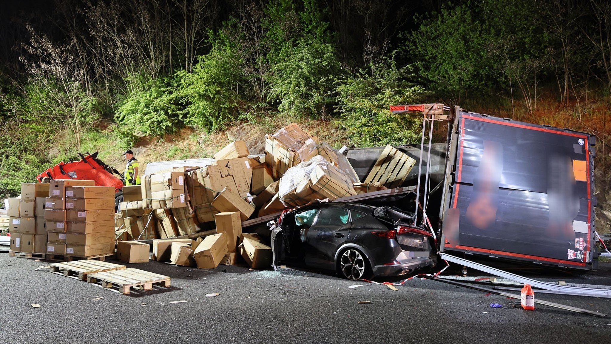 Umgestürzter Lkw auf der Autobahn, Kartons und Paletten liegen verstreut. Ein beschädigtes Auto ist unter der Ladung eingeklemmt. Einsatzkräfte sichern die nächtliche Unfallstelle.