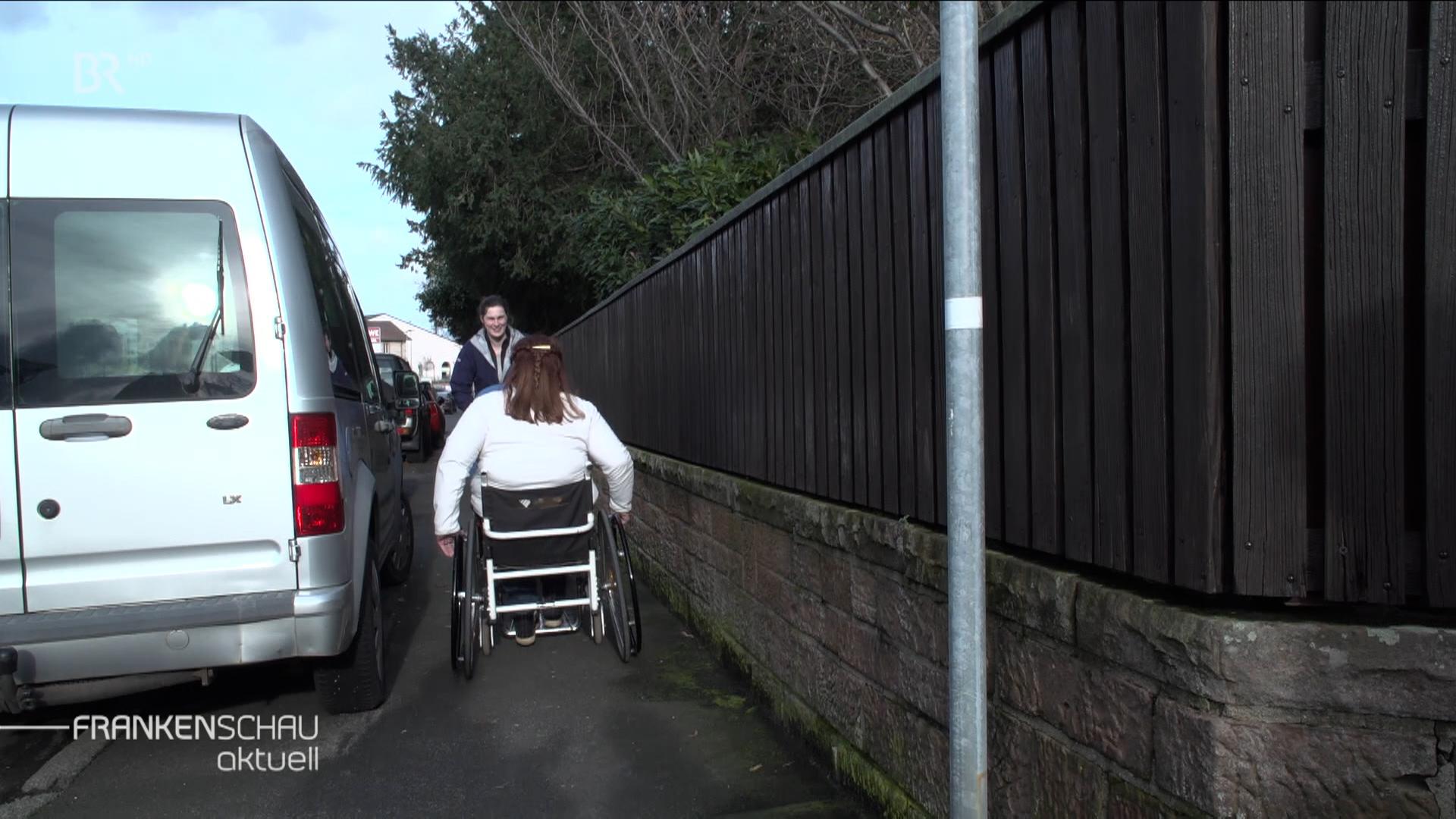 Eine Rollstuhlfahrerin fährt einer anderen Frau in einem engen Gehsteig entgegen.