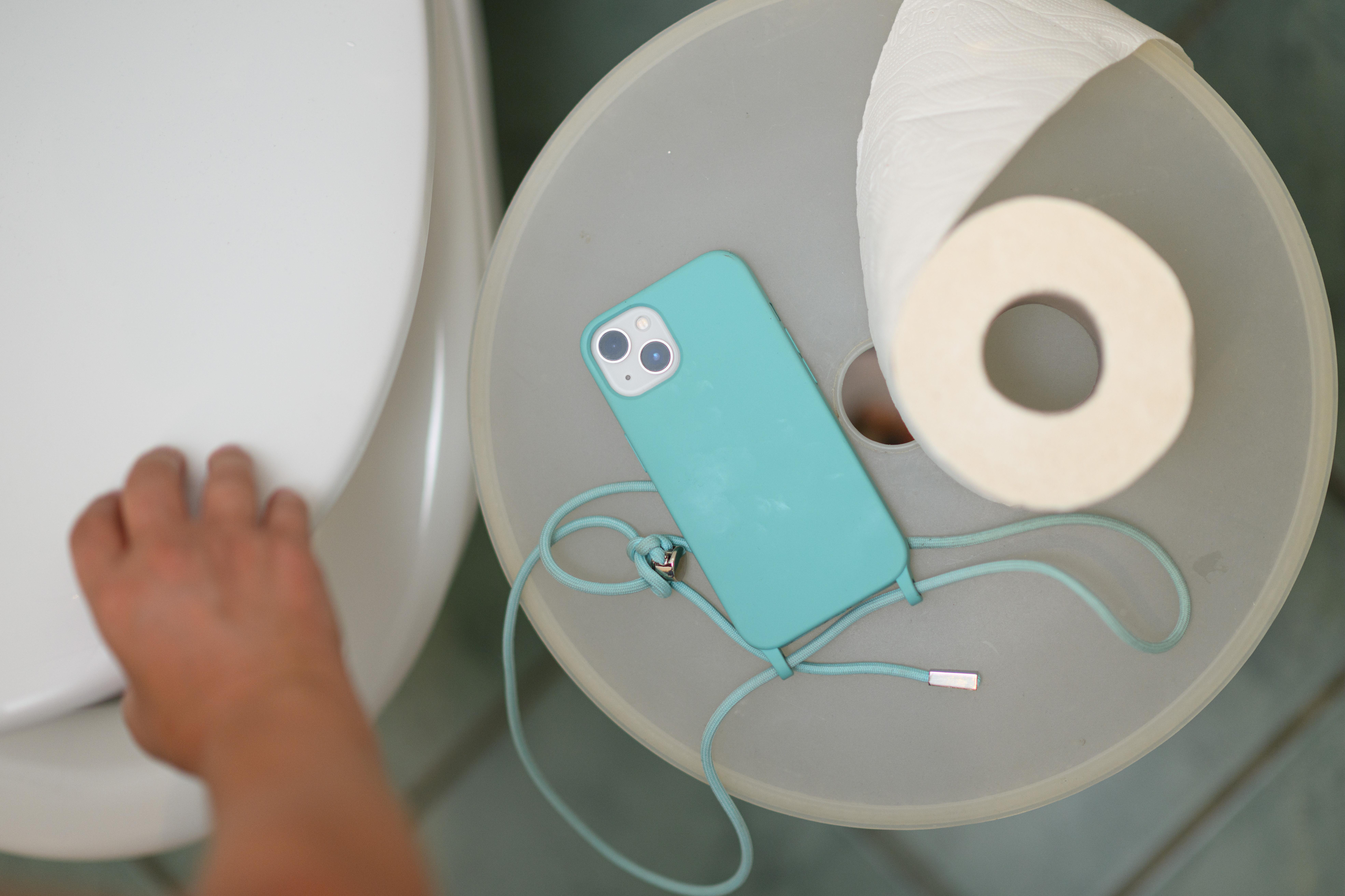 Ein Smartphone und eine Klopapierrolle liegen neben einer Toilette auf einem Mülleimer.