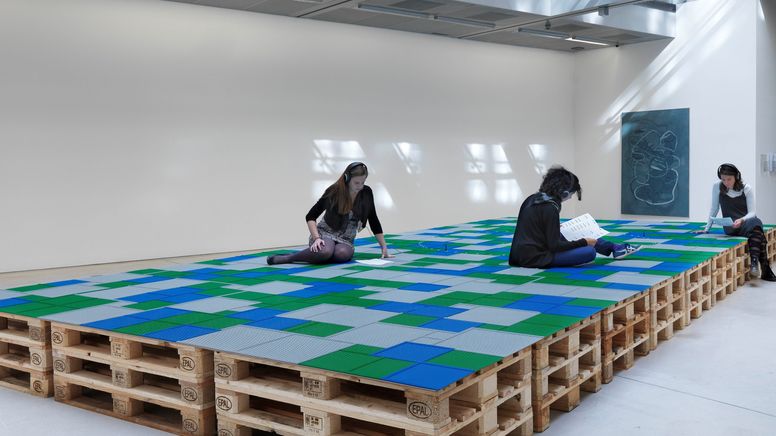 Drei Reihen übereinander gestapelter Europaletten, darüber blaue, grüne und graue Lego-Platten und junge Menschen, die darauf sitzen und jeweils ein Infoblatt lesen. | Bild:Courtesy die Künstlerin, Foto: Günter Kresser