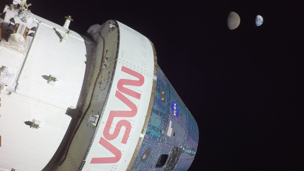 Im Vordergrund des Bildes befindet sich links die Raumkapsel Orion, die von außen sichtbar ist und ausschaut wie ein Kreisel; darunter befindet sich das European Service Module, das zylinderförmig ist. Rechts oben im Bild ist der graue Mond im Halbschatten zu sehen, kleiner rechts dahinter die Erde, ebenfalls im Halbschatten. 