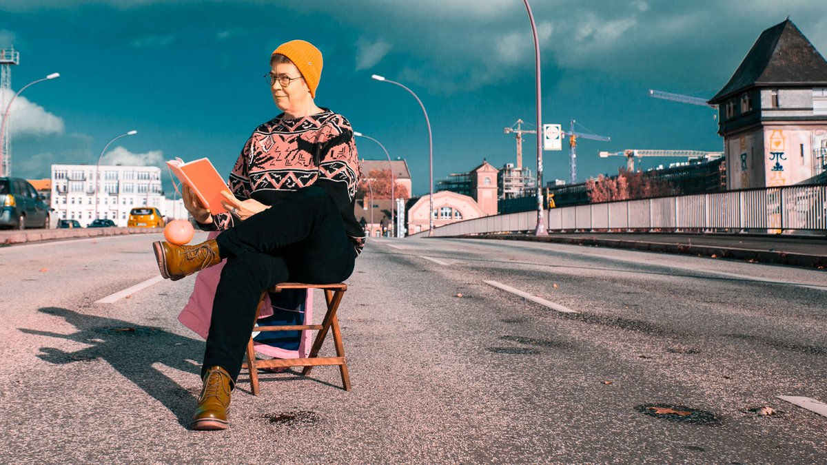 Die Philosophin Antke Engel, lesend auf einem Holzstuhl und auf einer Brücke mit Autoverkehr, bei sonnigem Wetteruer Himmel   