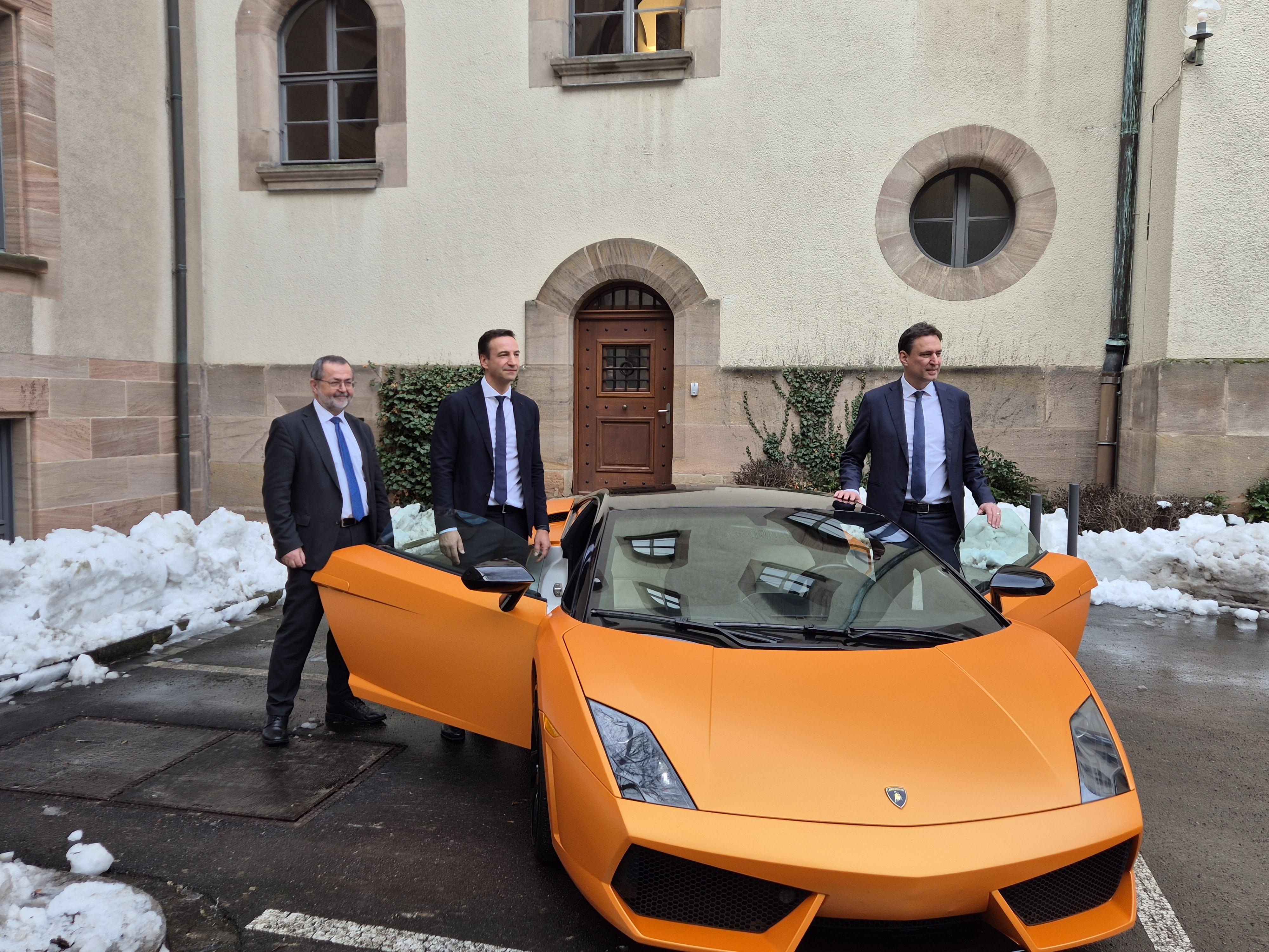 Drei Männer stehen um einen orangefarbenen Sportwagen. 