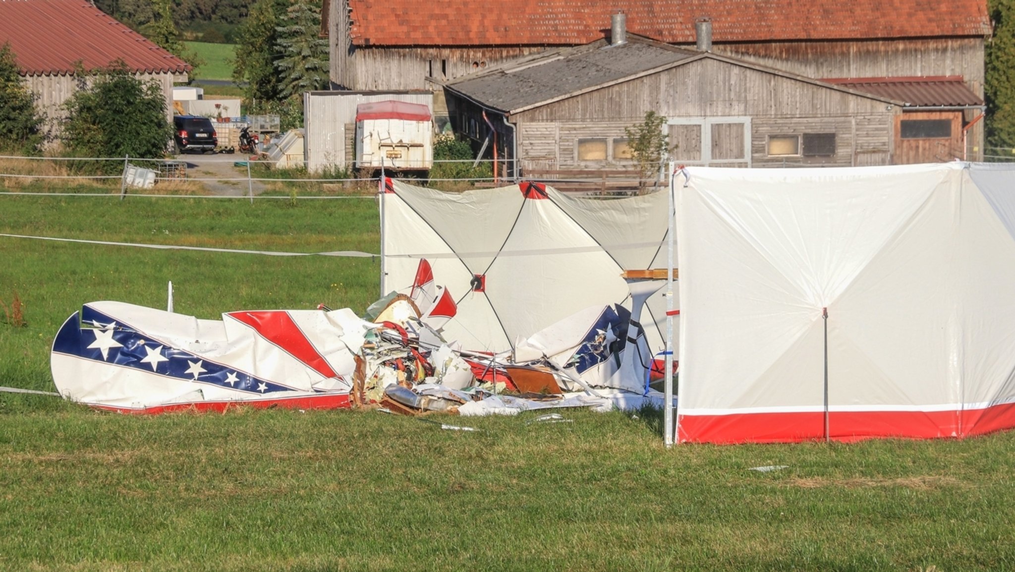 Auf einer Wiese liegen Teile des abgestützten Doppeldeckers, zum Teil abgeschirmt durch aufgestellte Sichtschutzwände.
