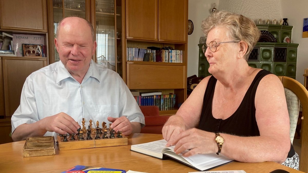 Ludwig Zier sitzt vor seinem Blinden-Schachspiel, neben ihm blättert seine Lebensgefährtin in einem Buch.