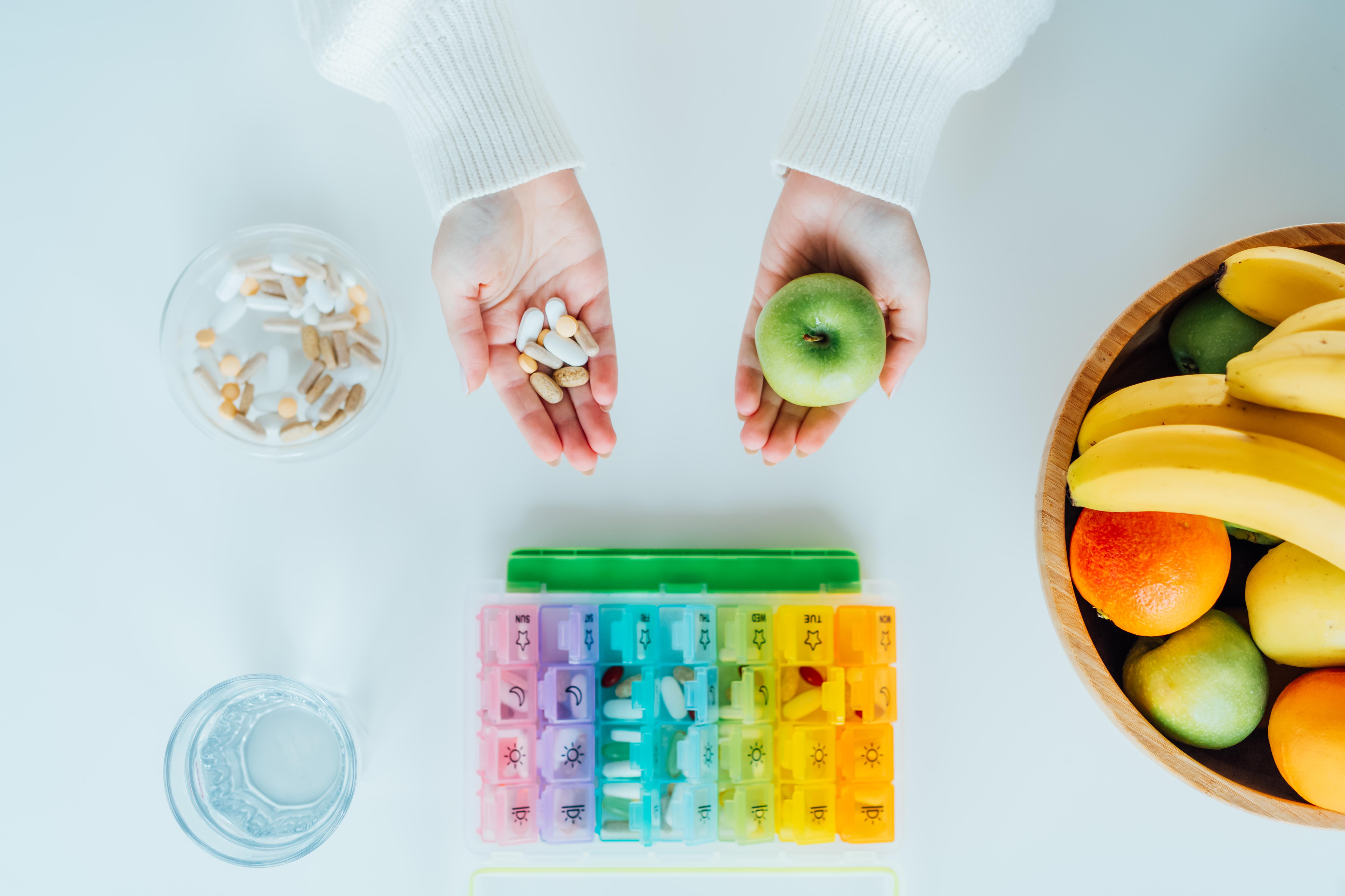Zwei Hände - jeweils mit Tabletten und einem Apfel, Pillenschachtel und Obstteller