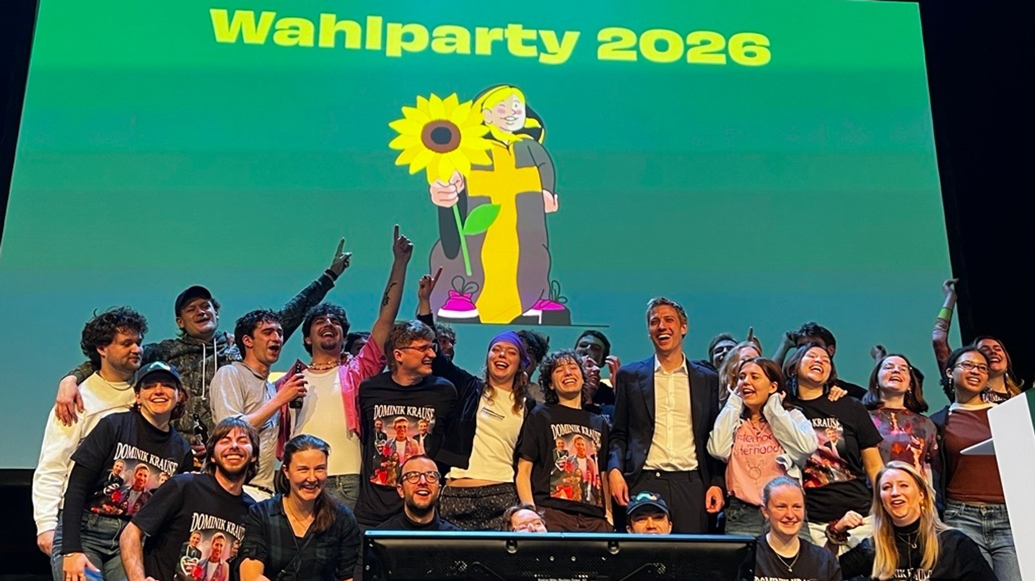 Dominik Krause und sein Wahlkampfteam auf der Bühne, alle freuen sich. Im Hintergrund eine wandfüllende Illustration des Münchner Kindls mit Sonnenblume vor grünem Hintergrund.