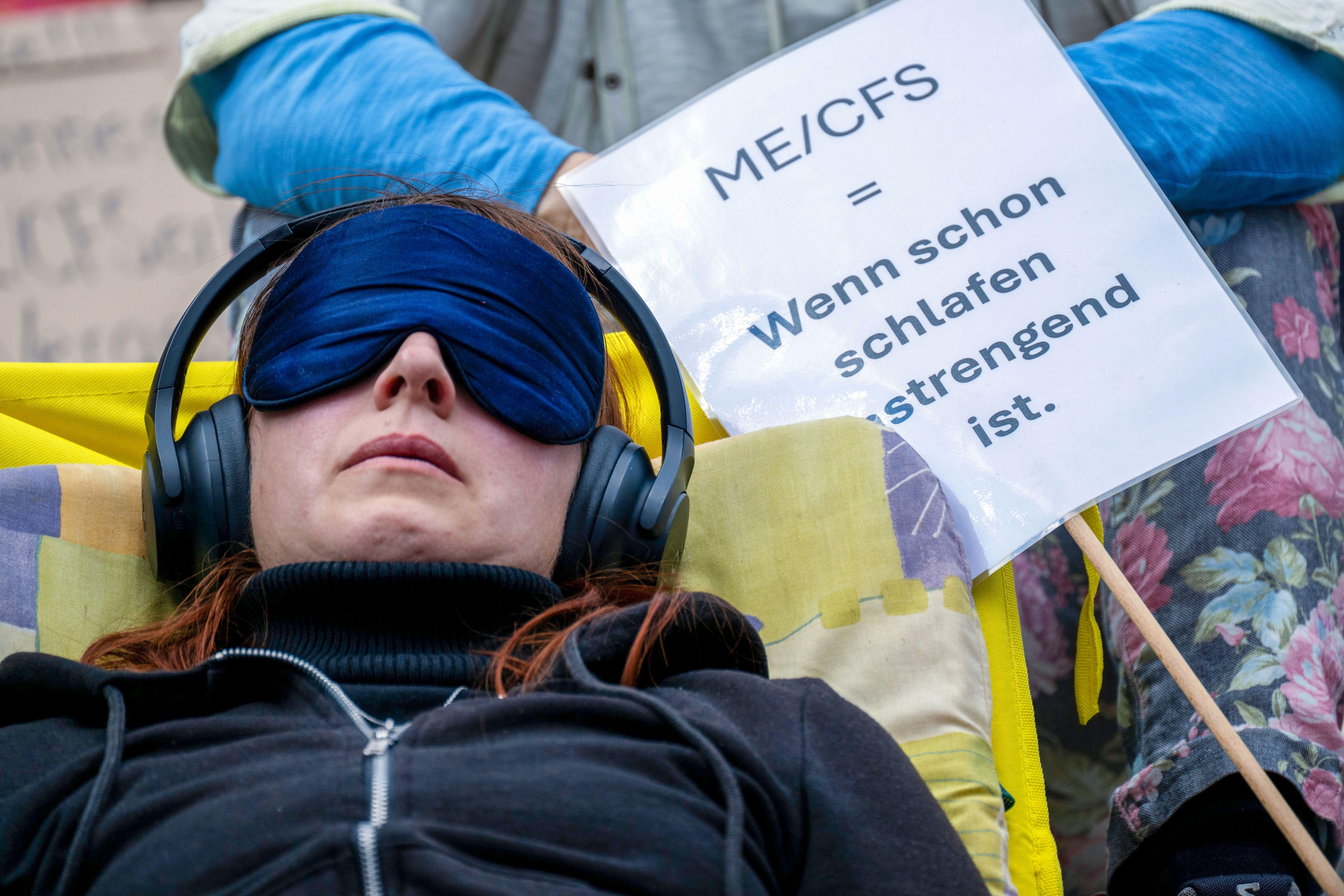 09.05.2026, Bayern, Nürnberg: Eine Frau liegt mit einer Schlafmaske und Kopfhörern auf einer Liege neben einem Schild mit der Aufschrift «ME/CFS = Wenn schon schlafen anstrengend ist.». Zum «Internationalen ME/CFS Awareness Day» machen Betroffene, Angehörige und Unterstützende auf die Situation von Menschen mit ME/CFS aufmerksam, einer schweren, bislang wenig erforschten Multisystemerkrankung. In Nürnberg beteiligen sich rund 400 Menschen an der Demonstration.