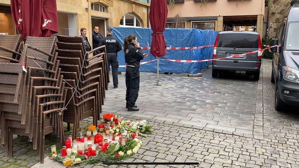 Polizeibeamte stehen vor dem abgesperrten Blumenladen. Auf dem Boden stehen Kerzen, Blumen und Grablichter.
